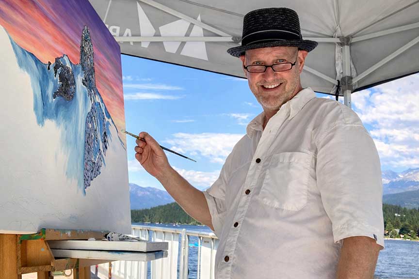 Artist graham watts painting a scenic landscape with mountains and water, standing outdoors under a tent. whistler bc