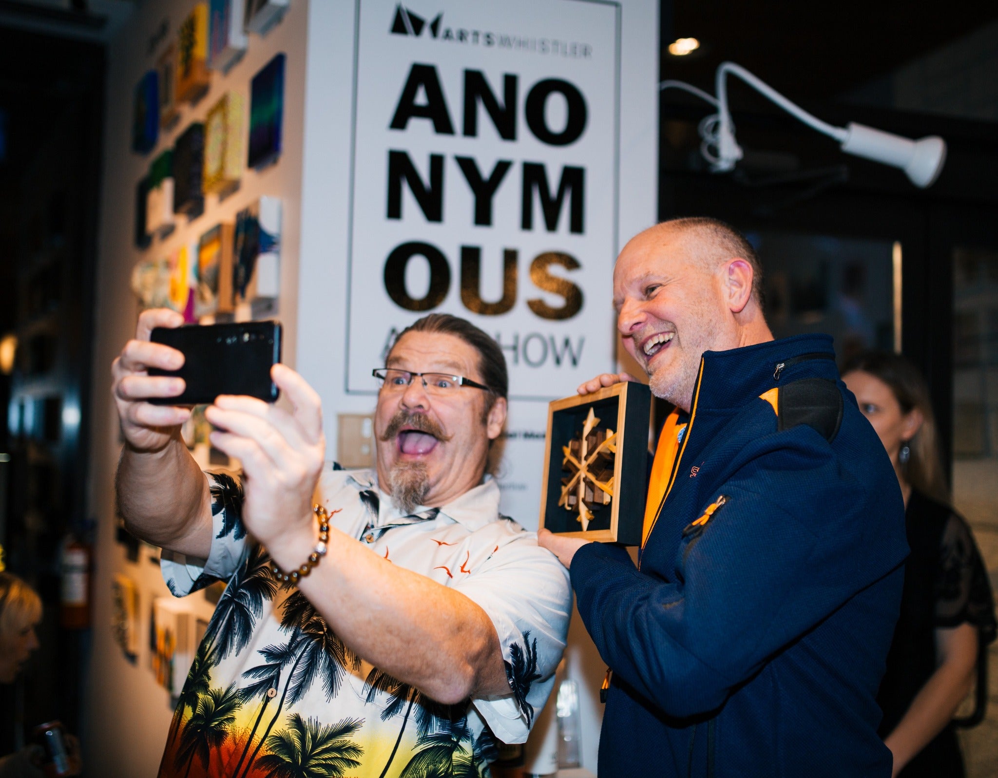 Two men taking a selfie with a phone, one holding a box, in front of a 'Anonymous Art Show in Whistler.  artist graham watts   