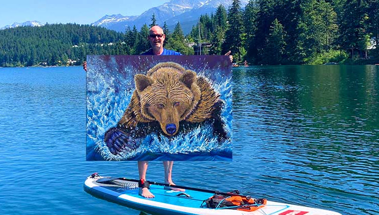Graham Watts on a paddleboard in Whistler BC holding a his painting bear-themed towel with a scenic lake and mountains in the background.