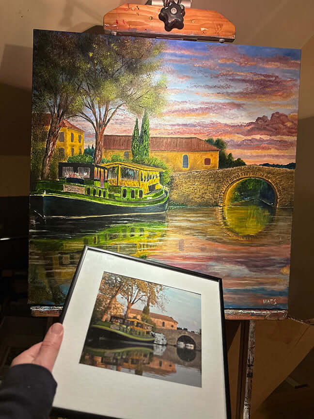 Person holding a framed print of a scenic painting with a boat on a river. fine art commission by artist graham watts 