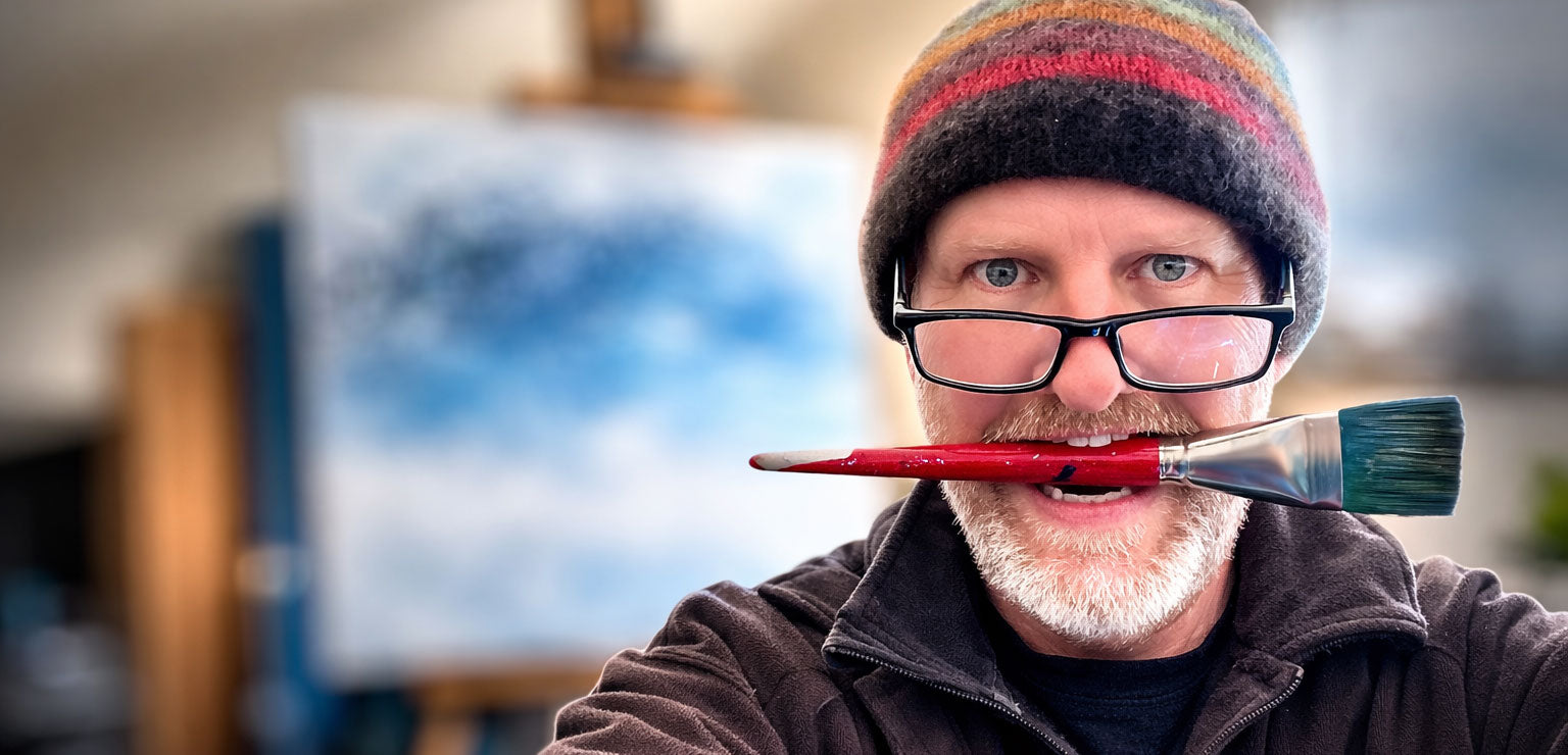 artist graham watts holding a paintbrush with a colorful hat and glasses, in front of an easel with a painting at his studio in vancouver, bc