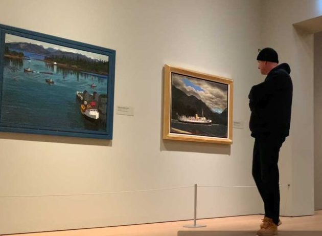 Photograph of the E.J. Hughes collection exhibit at the Audain Art Museum. The gallery features a diverse array of Hughes' artworks, including coastal landscapes and village scenes, showcasing his mastery of watercolor and oil painting techniques. Visitors explore the vibrant colors and intricate details of his iconic Canadian artwork.