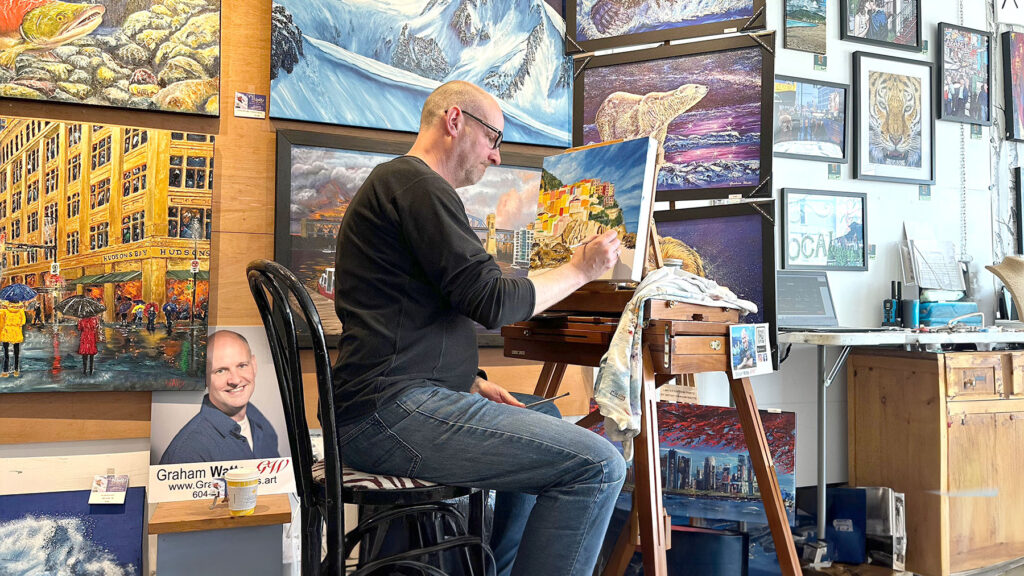 Artist Graham Watts working on a canvas in his art studio, surrounded by various vibrant paintings, including cityscapes and wildlife. A business card with contact information is visible on display.