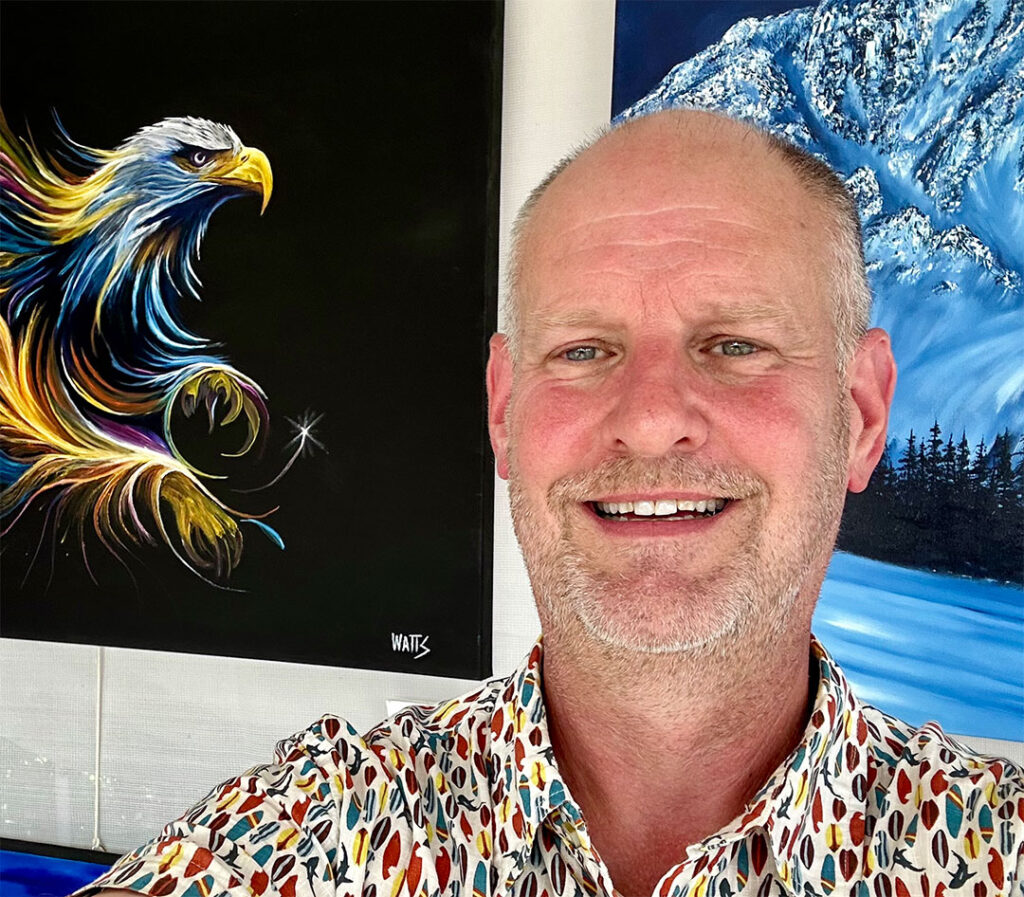 Artist Graham Watts smiling in his studio with a colorful, stylized eagle painting and a mountainous landscape in the background.